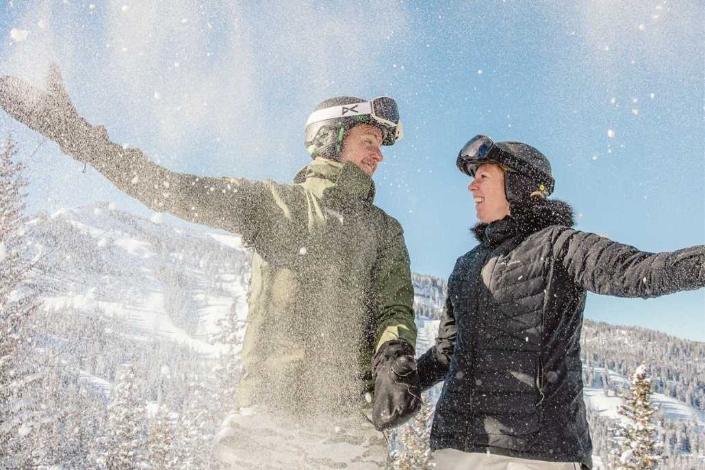 A couple dressed in winter gear and ski helmets joyfully throws snow into the air while standing on a snowy mountain in Aspen, with bright blue skies and snow-covered trees in the background.