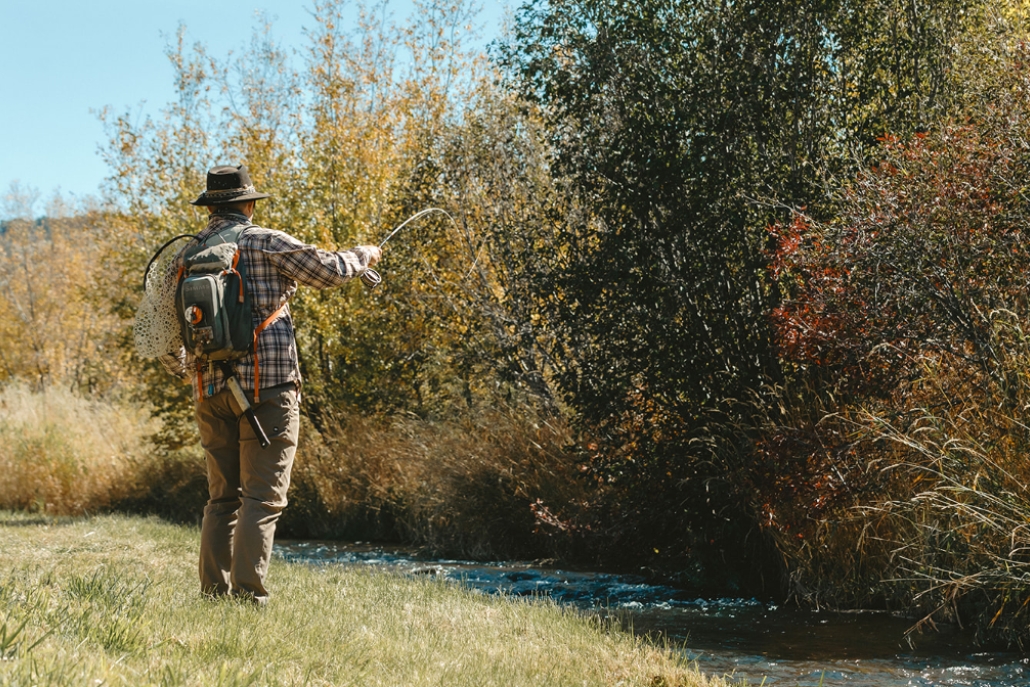 A man with a hat and backpack stands in grassy terrain beside a stream, framed by the fall colors of Aspen's Roaring Fork River.