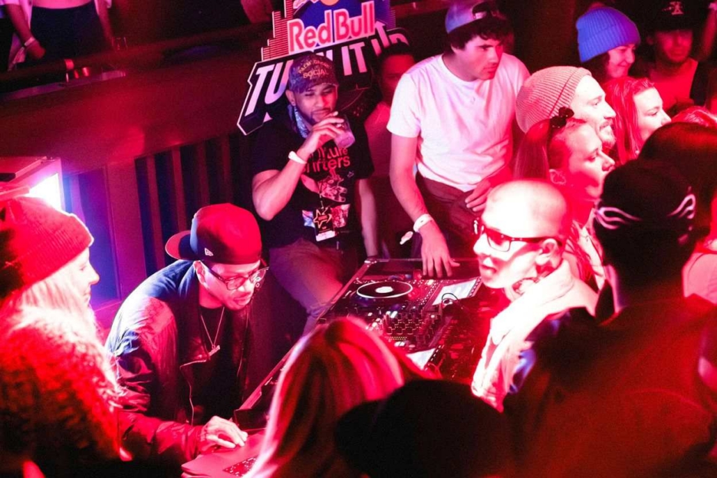 Crowd dancing near DJ booth under vibrant lights at a packed venue, energetic nightlife captured during a lively celebration.