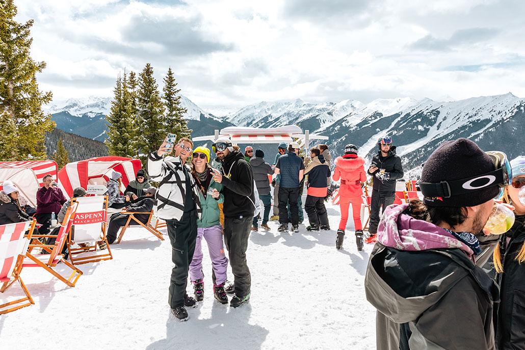 A group of four friends enjoying après-ski drinks, sitting outside on a sunny day with snow-covered mountains in the background.