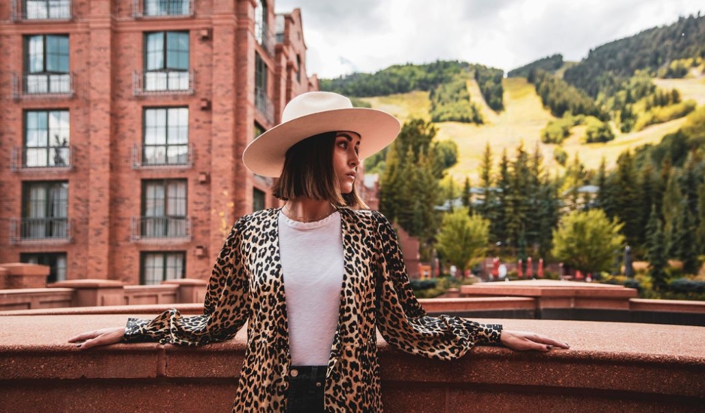 Stylish woman in a wide-brimmed hat and leopard print jacket standing near a red-brick hotel with Aspen Mountain in the background, capturing pre-winter travel vibes.