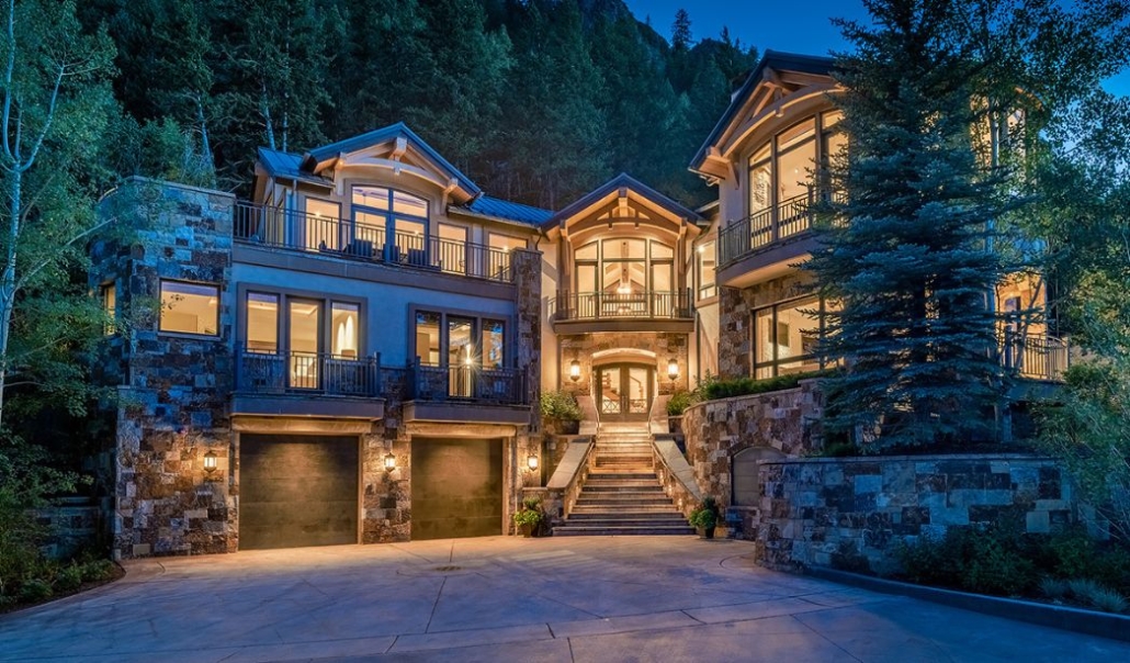 Exterior view of the luxurious Little Cloud Chalet in Downtown Aspen, featuring warm lighting, stone architecture, and forested mountain surroundings at twilight.