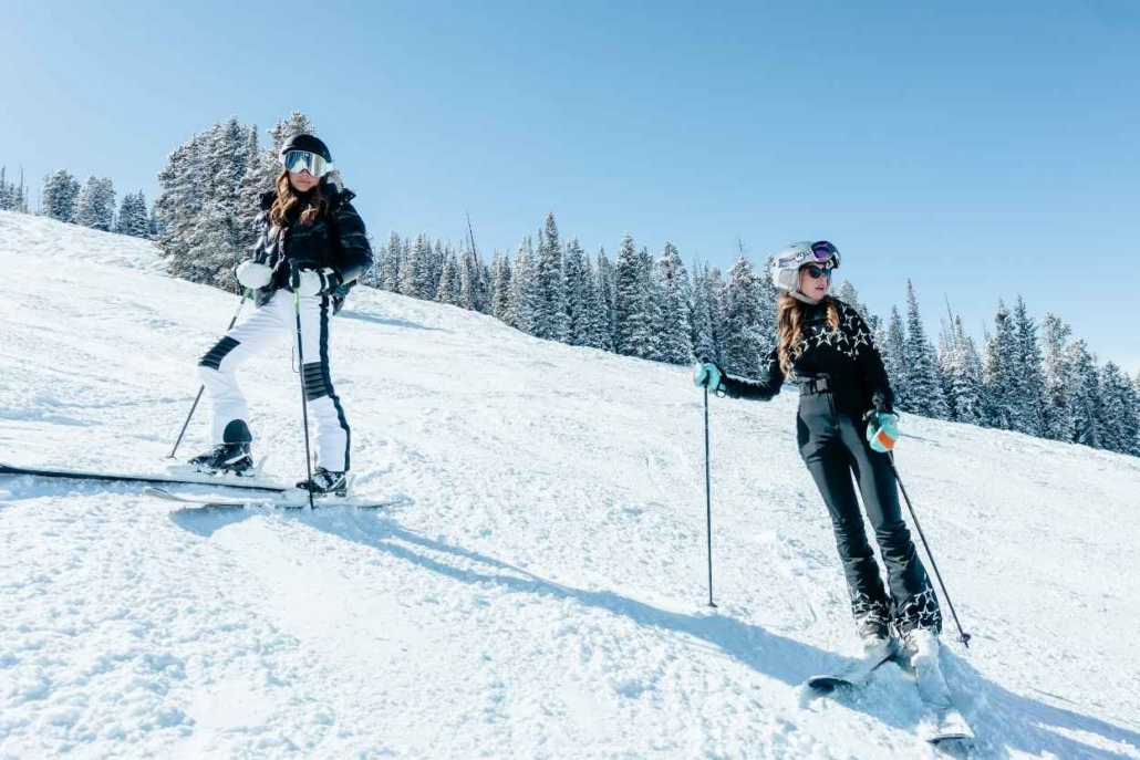 Two women in ski gear standing on a snowy slope with pine trees in the background, enjoying a sunny day on the mountain.