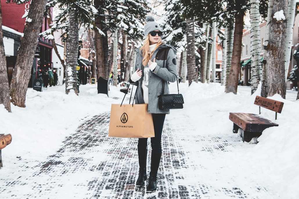 Stylish woman shopping in downtown Aspen during winter, carrying designer bags along a snowy, tree-lined street.