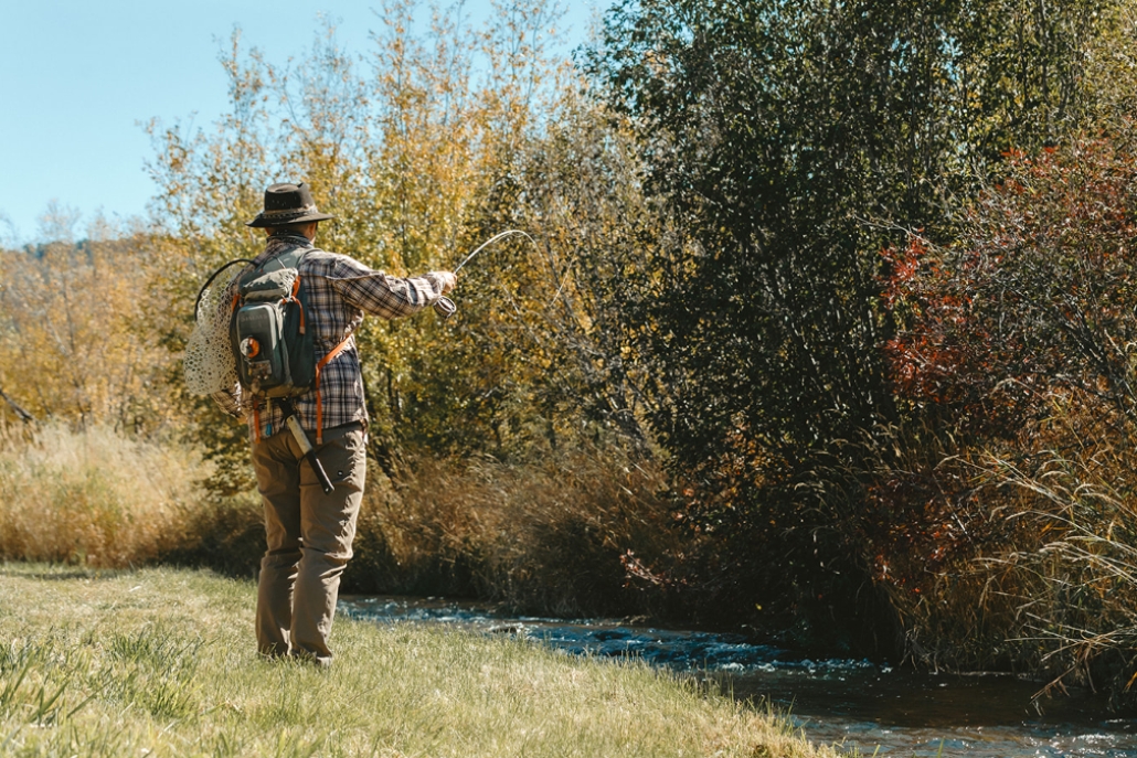 A man with a hat and backpack stands in a field beside a stream, showcasing Aspen's spring fly fishing scene.