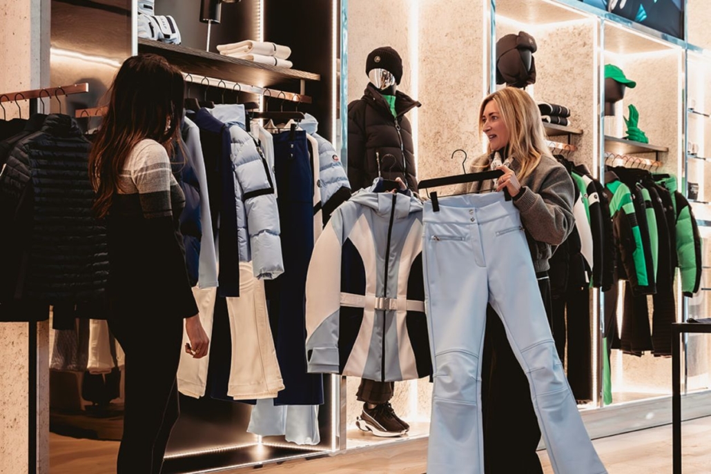 Two women browsing luxury winter wear at an upscale boutique in Aspen, featuring designer ski jackets, pants, and après-ski fashion items.