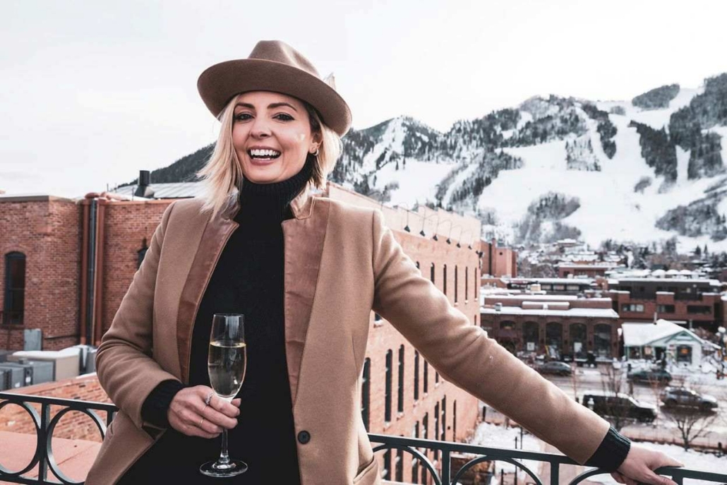 Smiling woman holding a glass of champagne on a rooftop in Aspen, with snow-covered mountains and red-brick buildings in the background.