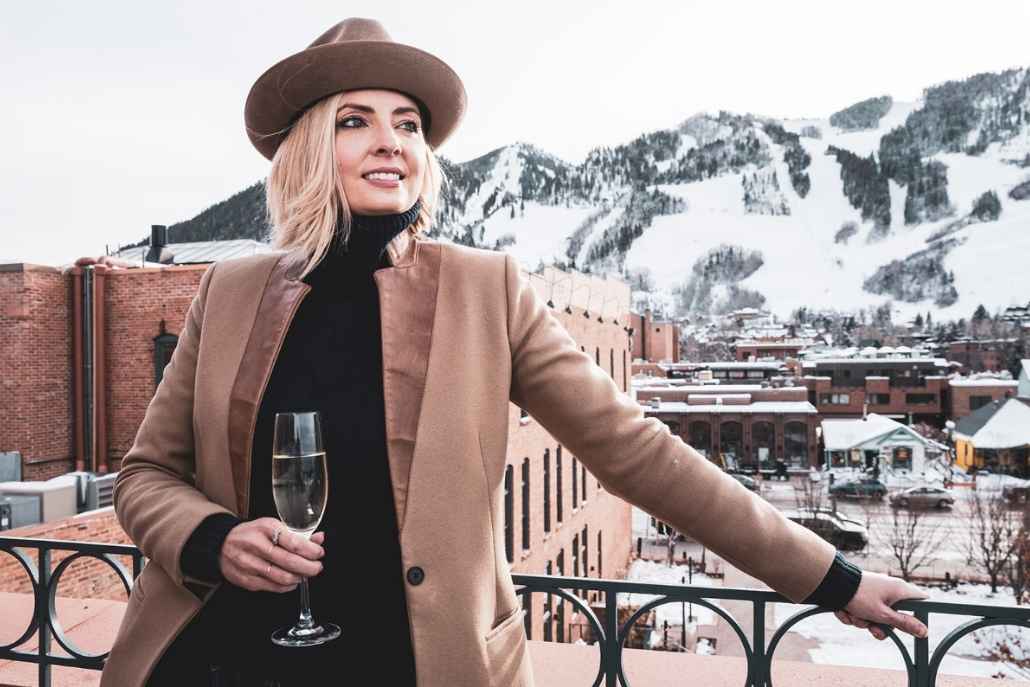 A woman wearing a hat and coat holds a glass of wine on a balcony, overlooking mountains during her Aspen Day 1 exploration.