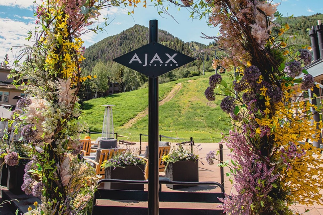 Entrance to Ajax Tavern in Aspen, framed by beautiful flowers and scenic mountain views, with ski lift visible in the background.