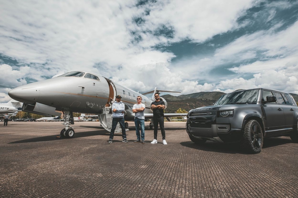 Three men standing in front of a private jet and a luxury SUV, with a scenic mountain backdrop, highlighting high-end travel to Aspen.
