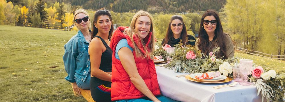 Group of five women sitting outdoors at a beautifully set table in Aspen, Colorado, surrounded by colorful autumn trees, as they enjoy their luxury trip.
