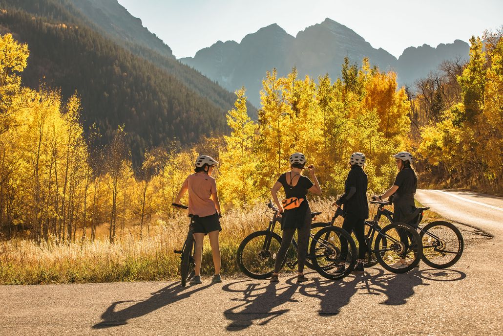 Four cyclists pause on a scenic Aspen trail, surrounded by golden fall trees and mountain peaks in the background.