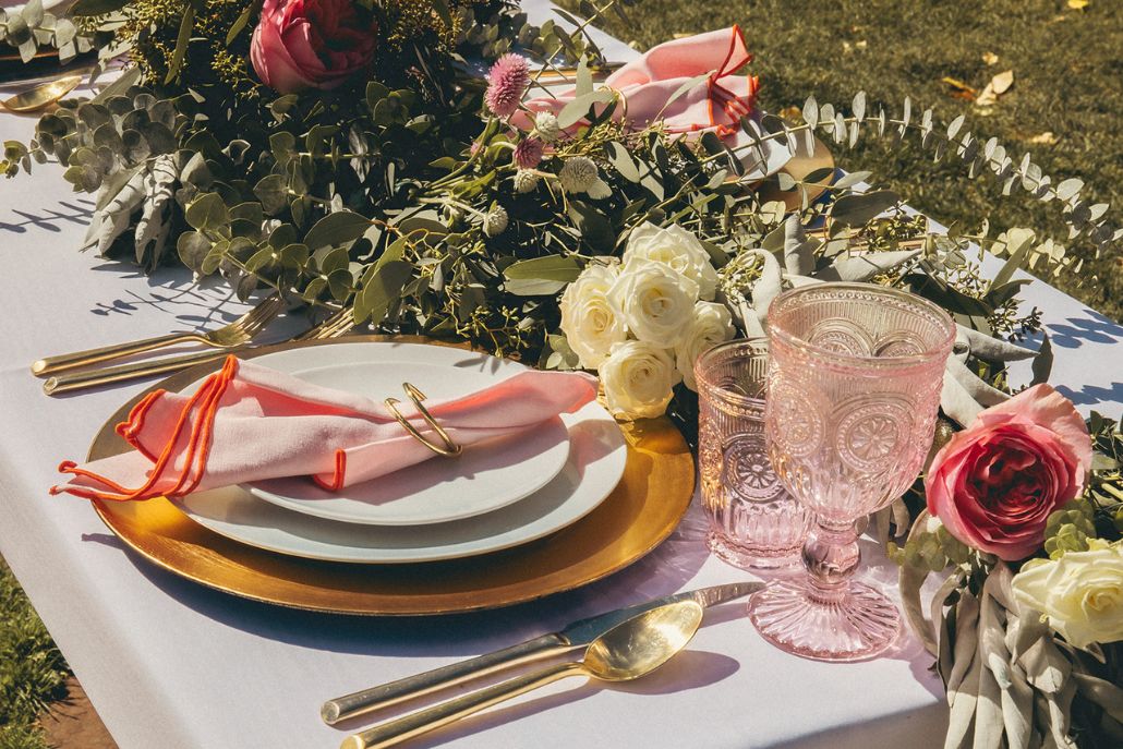 Elegant outdoor picnic setup with flowers, gold tableware, and pink glassware arranged on a table in a grassy setting.