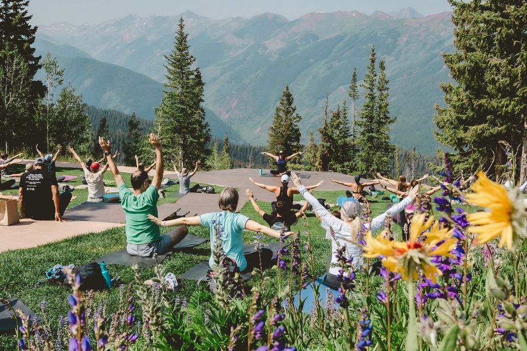 Group yoga session outdoors in the mountains, people stretching on mats with forested hills and wildflowers around.
